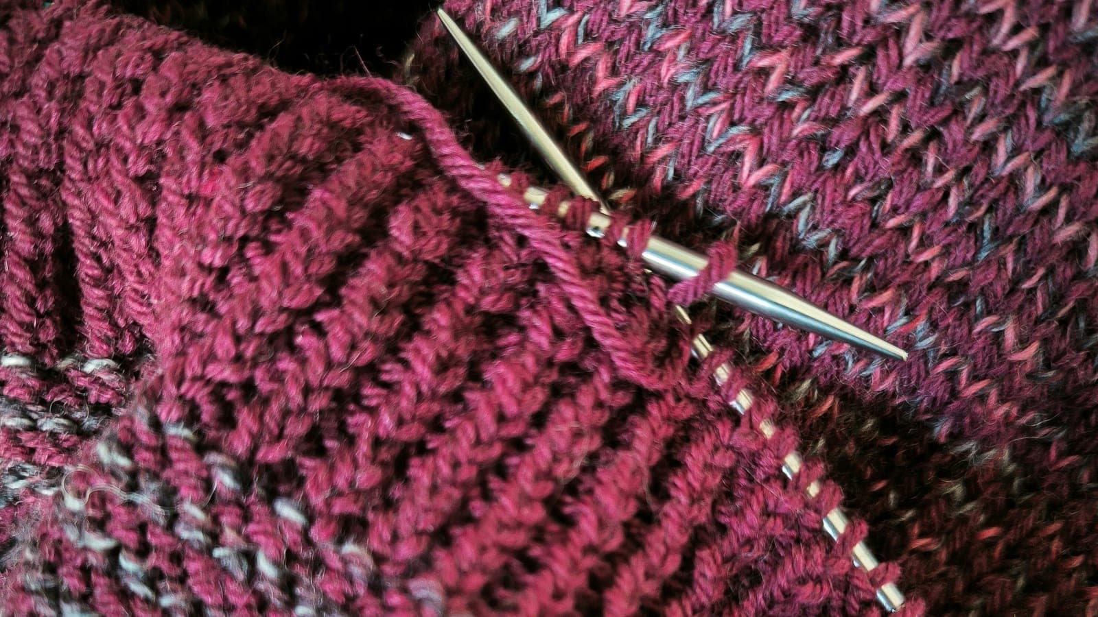 A hand-knitted piece in deep wine wool left mid-row, with a metal needle still holding the live stitches beside a finished ribbed edge.